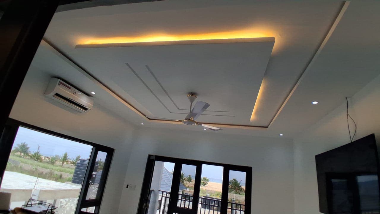 Open-plan living room in Keta with a stepped POP tray ceiling, warm cove lighting, and glass doors opening to the beachfront